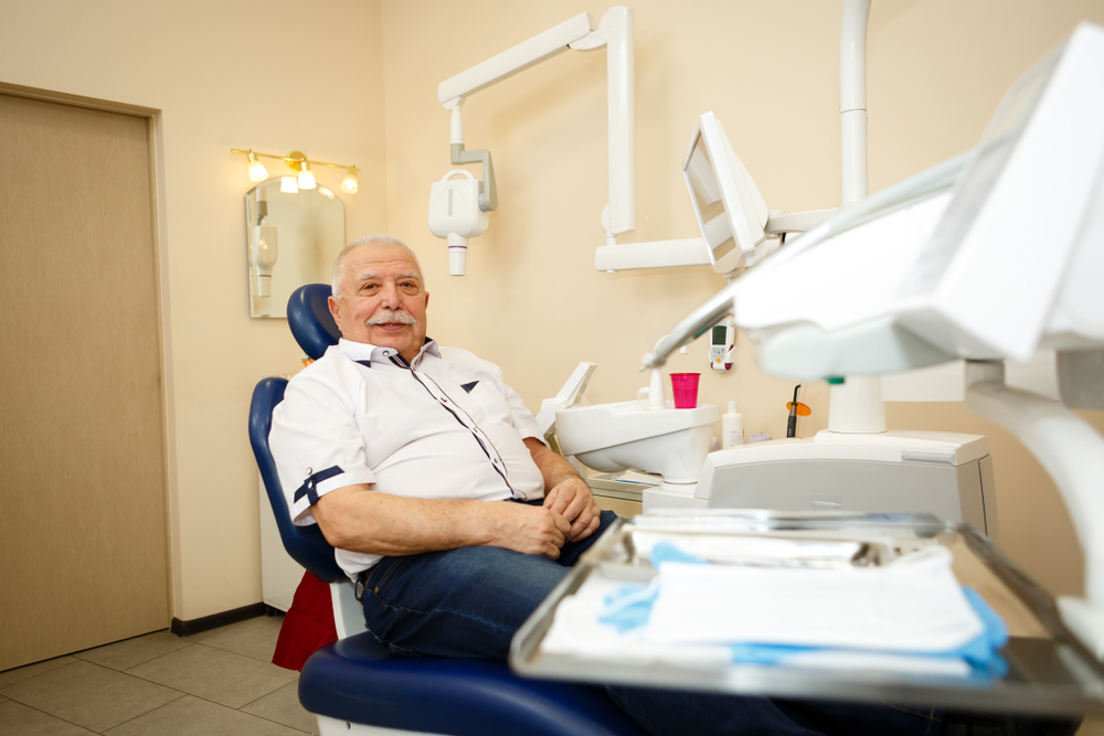 An older man with a white mustache sits in a dental chair, smiling. Dental equipment and tools are nearby. The background has beige walls, a mirror, and a closed door. The place looks like a dental clinic.