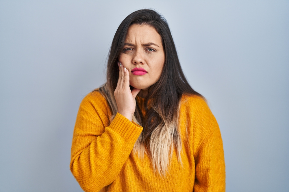 Woman with long dark hair and an orange sweater holds her cheek with a pained expression, suggesting toothache or facial pain, standing against a plain light blue background.