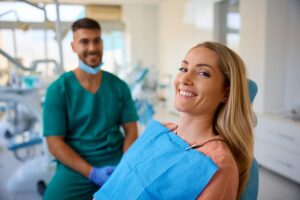 A woman smiles in a dental chair wearing a blue bib. A dentist in green scrubs and gloves sits nearby, also smiling. Dental equipment is visible in the background of the bright, modern clinic.