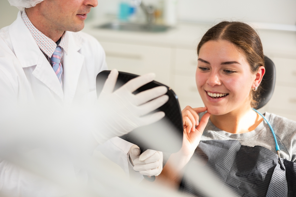 A woman sits in a dental chair, smiling and touching her chin as she looks at a handheld mirror. A dentist in a white coat and gloves stands beside her, holding the mirror. Dental instruments are visible in front.